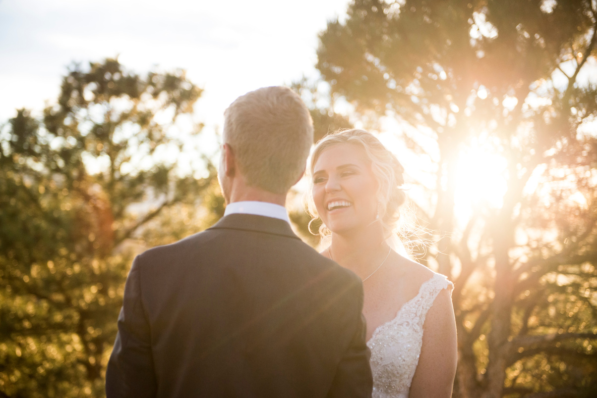 A bride smiles at her groom with the sun shining through the trees in the background.