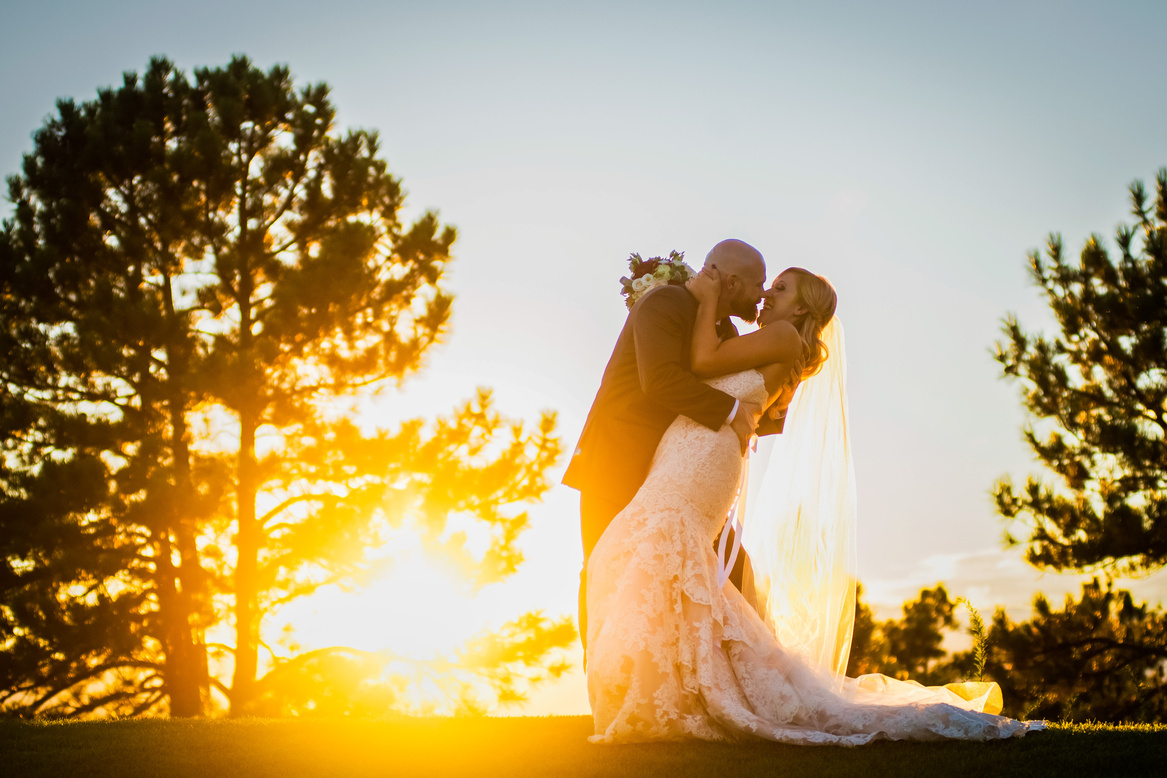 A bride and groom share a kiss with the sun shining behind them at sunset.