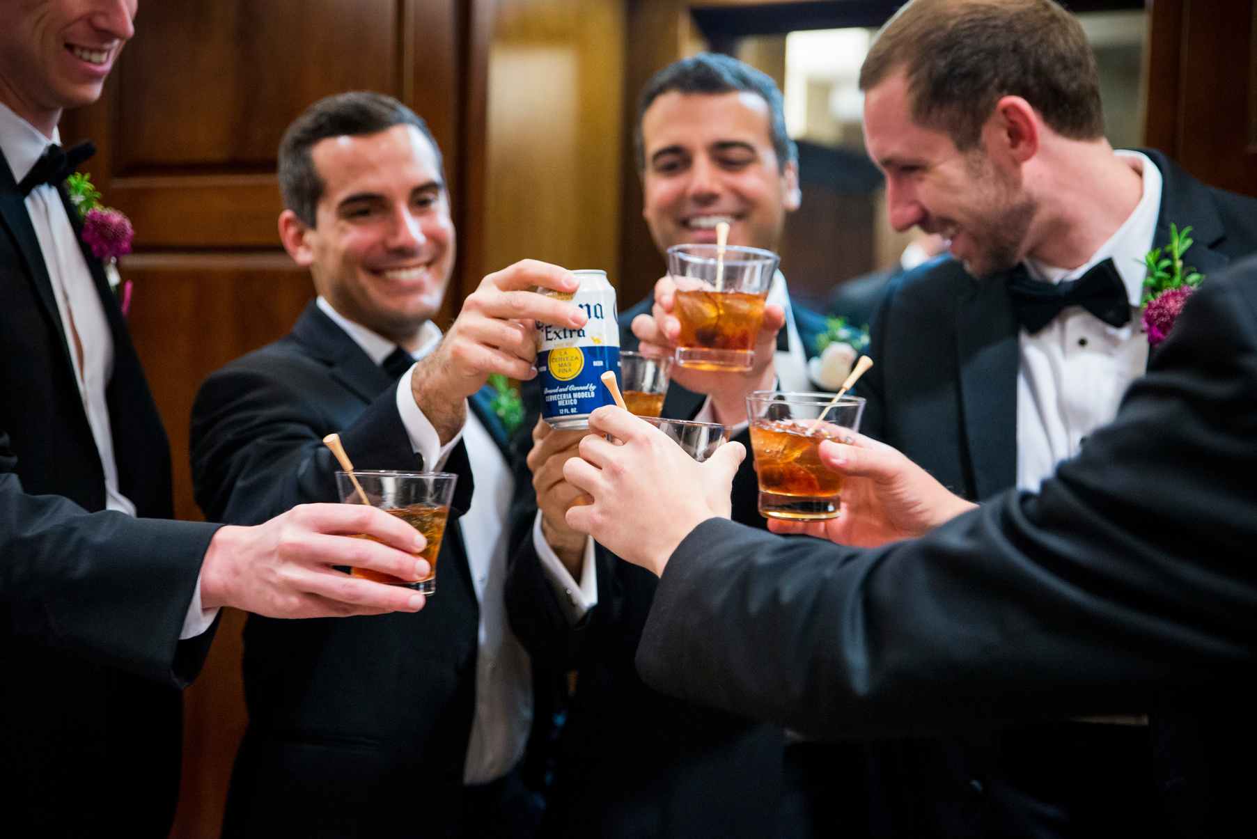 A group of groomsmen in tuxedos are toasting with beer and mixed drinks.
