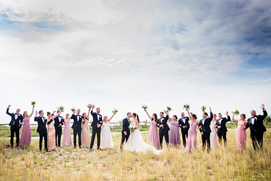 A group of bridesmaids and groomsmen pose for a photo in a field, throwing their hands in the air in celebration.