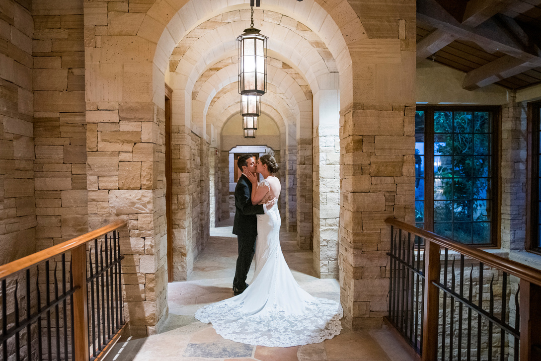 A bride and groom kissing in a stone hallway with arches.
