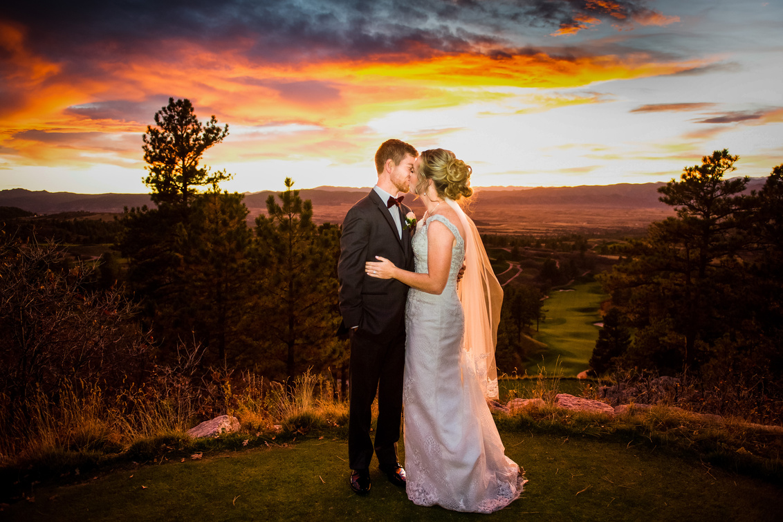 A bride and groom share a kiss on a mountain overlook at sunset with beautiful orange and pinks in the sky.