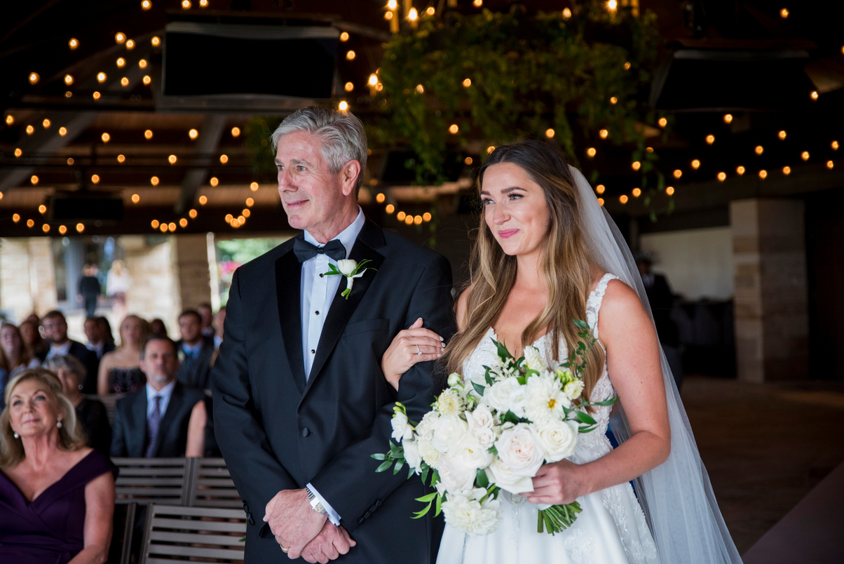 A bride and father walking down the aisle with twinkle lights in the background.